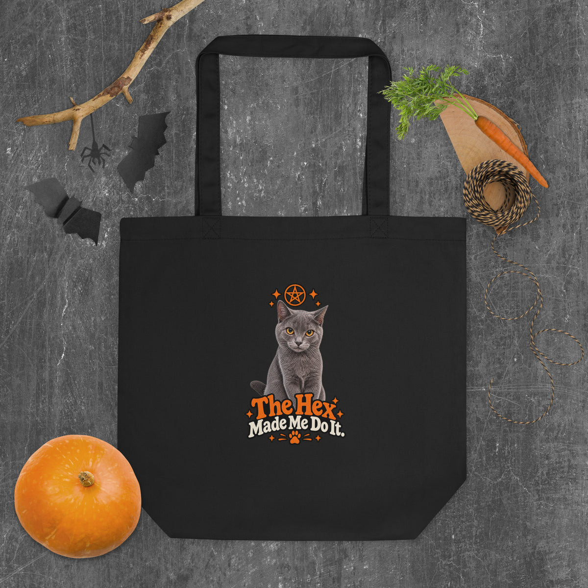 Black Uno the Cat Halloween tote bag featuring a gray cat, orange pentagram and stars, paw print, and “The Hex Made Me Do It” slogan, styled with pumpkins and Halloween decorations.