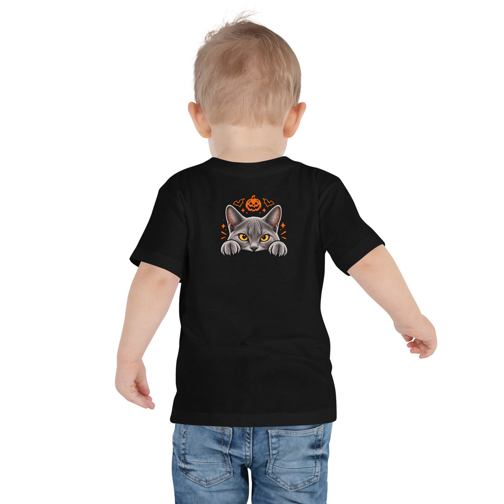 Back view of a toddler wearing a black Halloween tee with Uno the gray cat peeking over an edge, framed by an orange pumpkin and festive spooky accents.