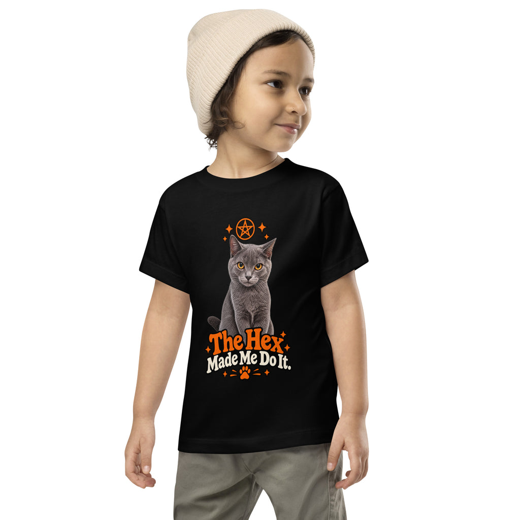 Child modeling a black toddler Halloween T-shirt with Uno the gray cat, orange mystical-style accents, and “The Hex Made Me Do It” lettering.